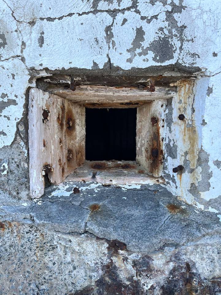 An old, weathered window frame with peeling paint on a textured wall, looking into a dark interior.