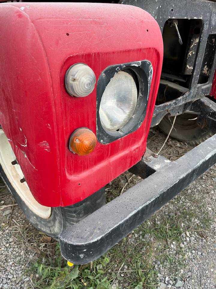 Close-up of the front corner of a red vintage vehicle showing a headlight, a turn signal, and part of a bumper against a gravel background. The vehicle is weathered and its paint is cracked.
