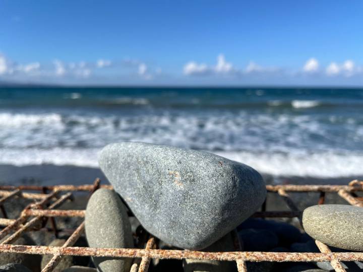 A close-up of smooth stones on a rusty metal grid with a beach and choppy ocean waves in the background.